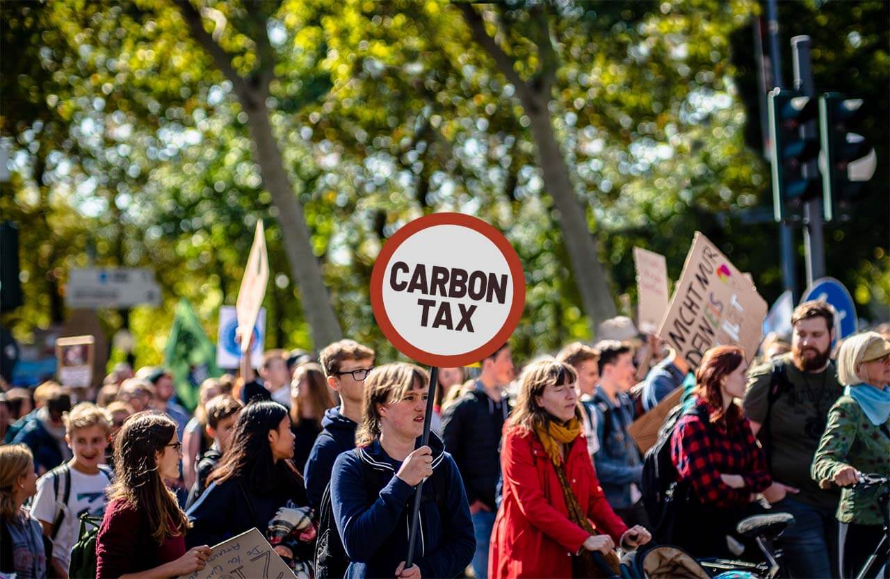 Pessoas numa passeata contra as mudanças climáticas carregando cartazes, dentre os quais destaca-se um que incentiva tributar o carbono.