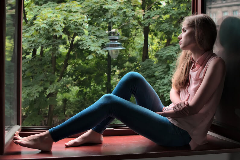 Foto de uma mulher jovem sentada na janela de uma casa, com olhar distante para o verde do lado de fora, ilustra o post sobre o que fazer se a procrastinação afetar sua saúde.