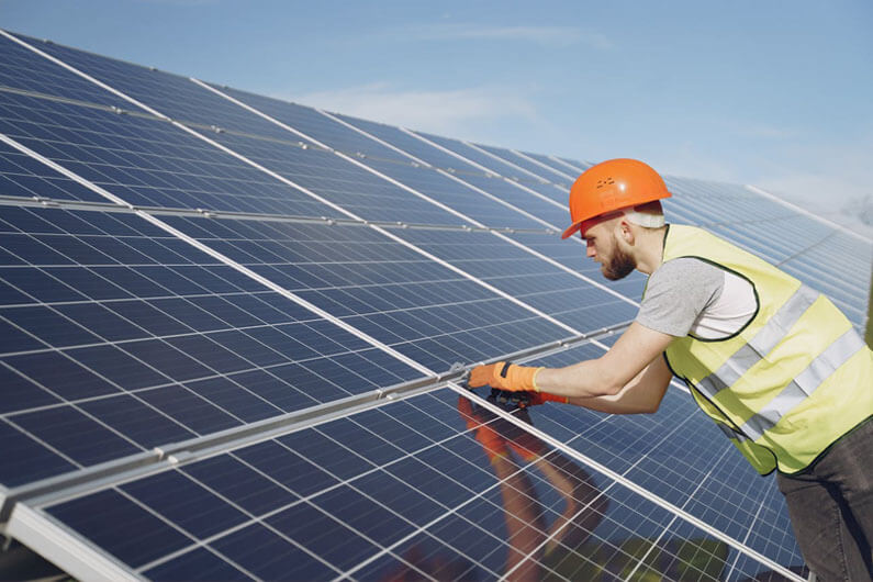 Imagem de um trabalhador usando luvas e capacete laranja instalando painéis solares no topo de um telhado ilustra o artigo cujo título diz que as energias eólica e solar são energias com crescimento histórico mais rápido.