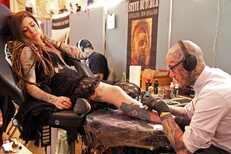 Imagem de um homem fazendo uma tatuagem na perna de uma mulher ilustra o post cujo título diz que as tatuagens aumentam o risco de câncer em 21 por cento.