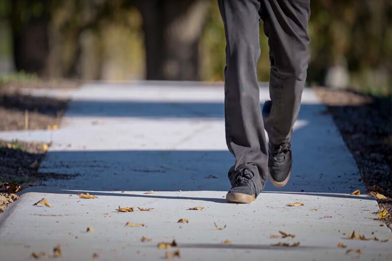 Imagem das pernas de uma pessoa vestindo calças cinzas e caminhando sobre uma calçada de concreto ilustra o post cujo título diz que um super concreto reforçado com café vai começar a ser testado.