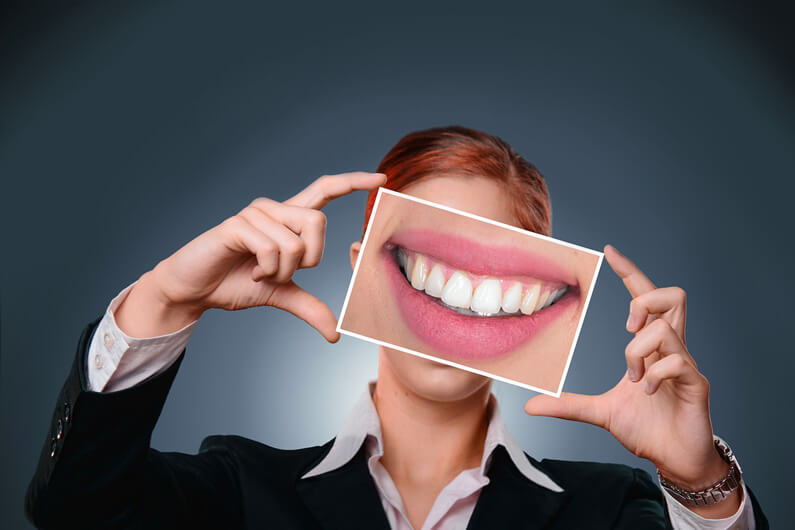 Imagem de uma mulher vestida de roupa executiva segurando na altura da cabeça uma foto de uma boca sorrindo com os destes à mostra ilustra o post cujo título diz que vai iniciar o teste do primeiro medicamento para regenerar dentes.