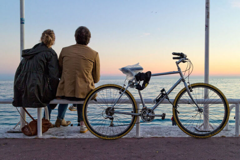 Imagem de uma bicicleta estacionada atrás de um casal que está sentado em frente ao mar e contemplando o pôr do sol ilustra o post cujo título diz que um medicamento antienvelhecimento prolonga a vida em até 25%