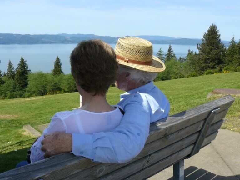 Imagem de um casal idoso sentado em um banco de madeira olhando para o mar ilustra o post cujo título diz que fatores de risco de demência podem reduzir em 45 por cento.