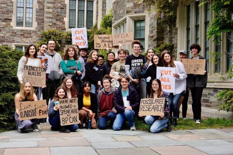 Imagem ao ar livre de alunos da Universidade de Princeton segurando cartazes de protestos à ligação entre a universidade e as empresas de combustíveis fósseis ilustra o post cujo título diz que as empresas de combustíveis fósseis estão sequestrando universidades.