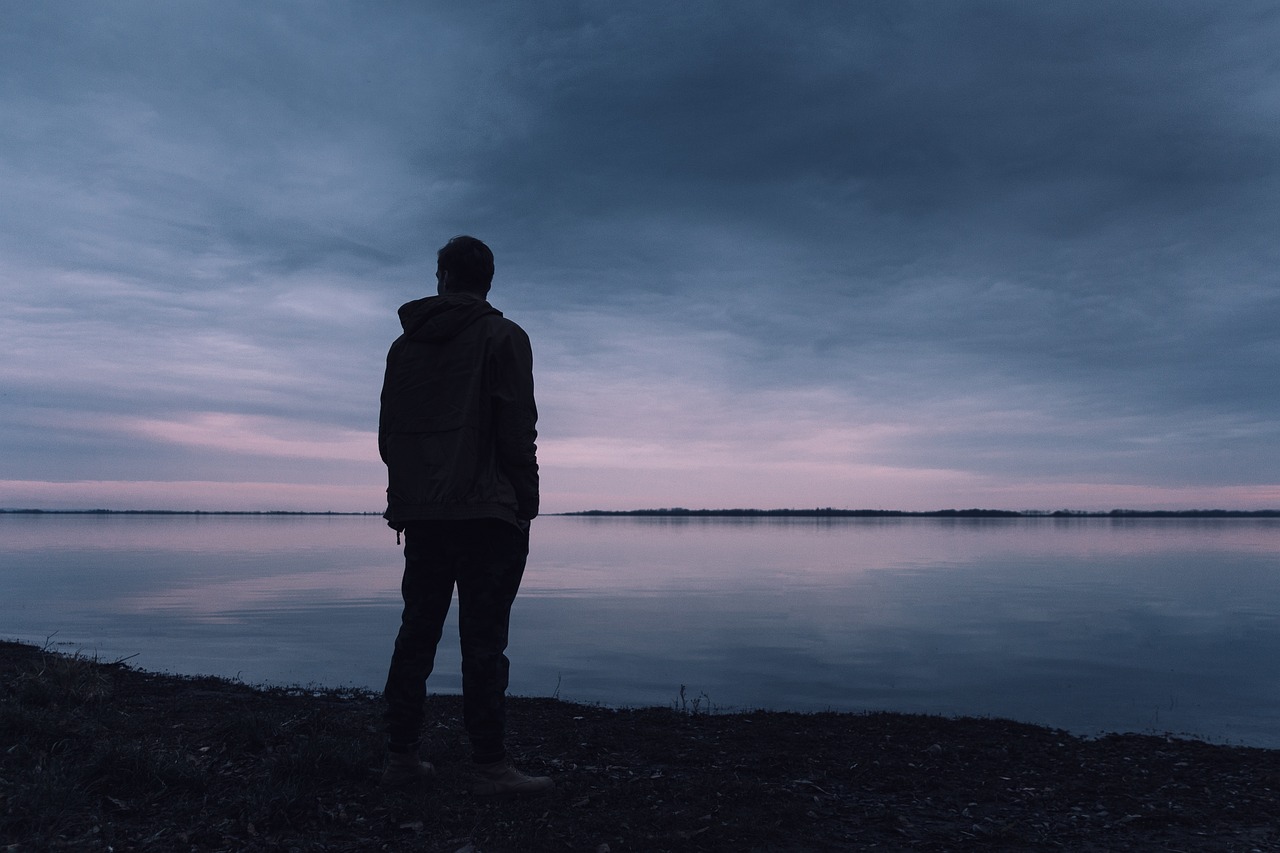 Imagem de um homem sozinho contemplando um lago num final de tarde sob um céu nublado e acinzentado ilustra o post cujo título diz que os homens estão carregando o peso da ‘epidemia de solidão’ em meio a potentes pressões sociais.