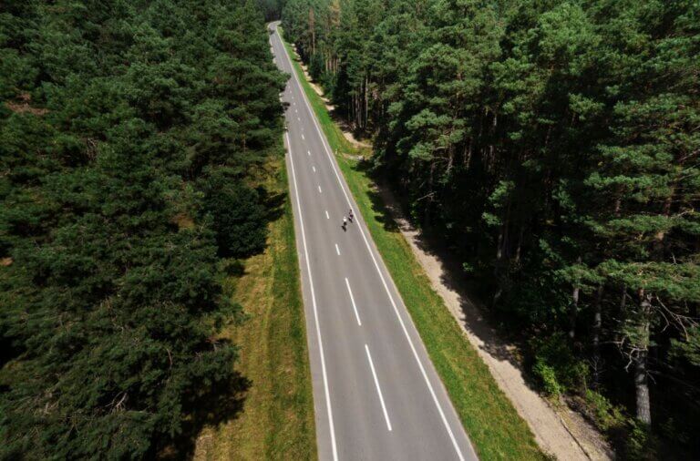 Imagem aérea de uma estrada cercada de uma floresta e duas ciclistas pedalando por ela ilustra o post cujo título diz que a neutralidade de carbono é uma estrada e não um destino.