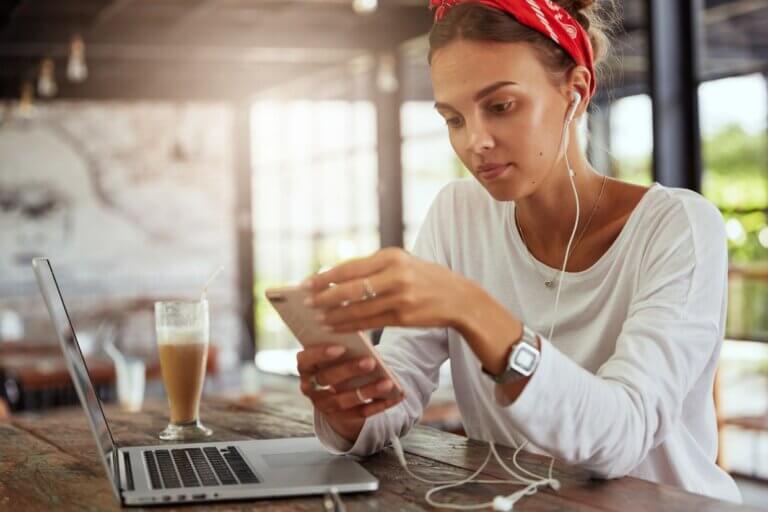 Imagem de uma mulher com fones de ouvido plugados em um celular junto a um laptop ilustra o post sobre como superar a dependência dos eletrônicos e fazer um detox digital de sucesso.