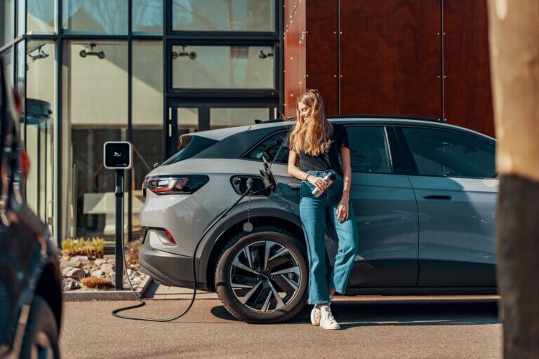 Imagem de uma moça recostada em um carro cinza que está sendo alimentado por uma tomada de eletricidade ilustra o artigo cujo título diz que os veículos elétricos não são verdadeiramente carbono zero.