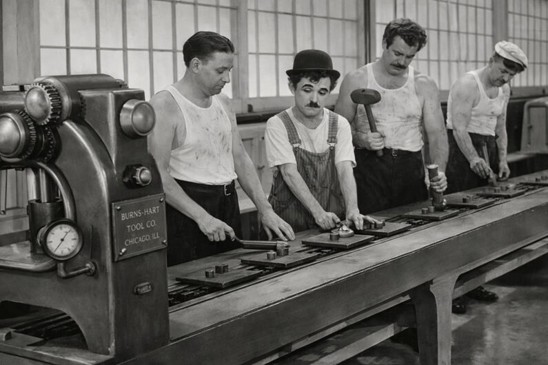 Imagem de Charles Chaplin apertando peças junto a três operários no filme Tempos Modernos ilustra o post que aborda por que a mão de obra global está desengajada, esgotada e doente.
