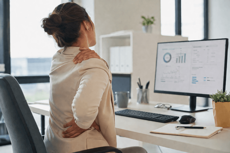 Imagem de uma mulher sentada na frente de um computador de mesa e com as mãos no ombro e na cintura ilustra o post que diz que sentar o dia todo pode estar destruindo sua saúde.