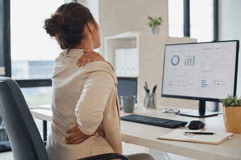 Imagem de uma mulher sentada na frente de um computador de mesa e com as mãos no ombro e na cintura ilustra o post que diz que sentar o dia todo pode estar destruindo sua saúde.