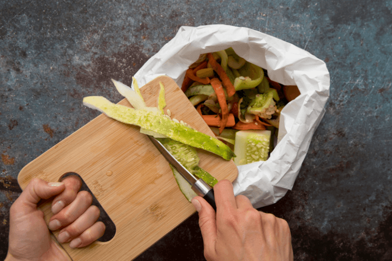 Imagem de uma mão segurando uma faca que raspa o alimento de uma tábua para dentro de uma lata de lixo para ilustrar por que os europeus desperdiçam tanto alimento.