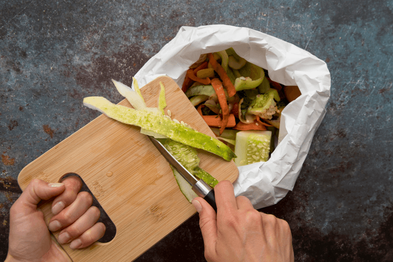 Imagem de uma mão segurando uma faca que raspa o alimento de uma tábua para dentro de uma lata de lixo para ilustrar por que os europeus desperdiçam tanto alimento.