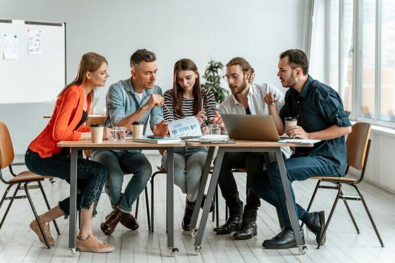 Imagem de três homens e duas mulheres reunidos em volta de uma mesa com um laptop ao lado de copos, e blocos de anotação ilustra o post cujo título diz que as empresas grandes apertam o freio nas metas de sustentabilidade.