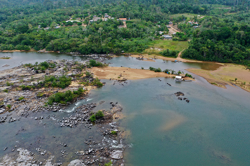 Imagem do leito do rio Xingu ao lado de uma área com algumas casas no meio de uma floresta ilustra o post sobre o dilema de Belo Monte e o conflito por água do rio Xingu.