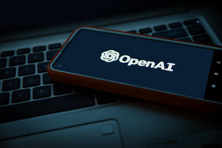 Imagem do nome OpenAI que aparece na tela de um celular que está sobre o teclado de um laptop ilustra o post cujo título diz que a OpenAI reduz desenvolvimento de IA avançada após ciberataque.