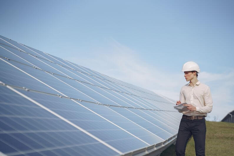 Imagem de um trabalhador usando um capacete branco e ao lado de placas solares ilustra o post cujo título diz os empreendimentos verdes em estágio inicial podem gerar bons empregos.