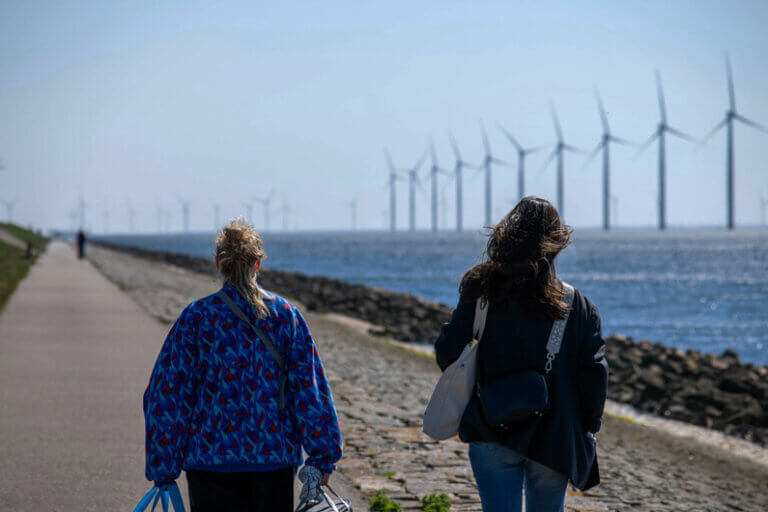 Imagem de duas mulheres caminhando à beira mar (ou de um lago) que mostra como pano de fundo várias turbinas eólicas offshore ilustra o post cujo título diz que o pêndulo da sustentabilidade não vai retornar para onde saiu.