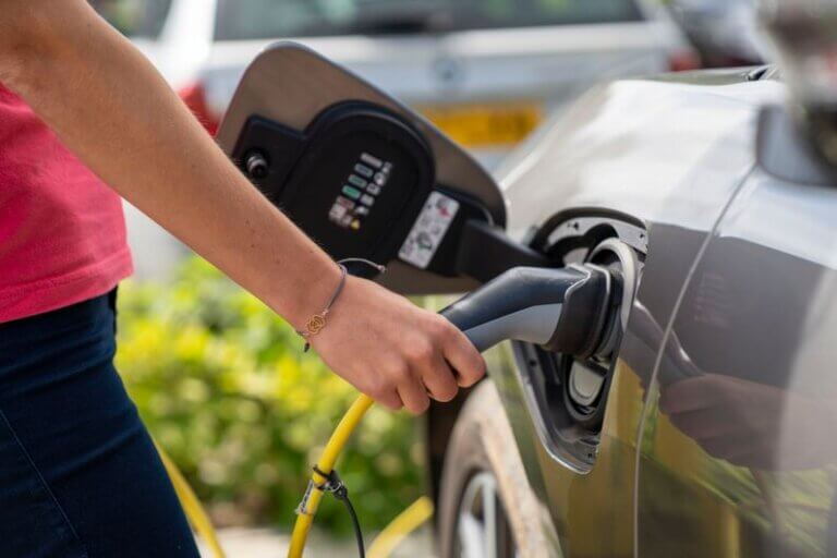 Imagem do braço de uma pessoa segurando uma tomada de carregamento acoplada a um EV ilustra o post cujo título diz que a crise de energia impulsiona a adoção de carros elétricos no mundo.