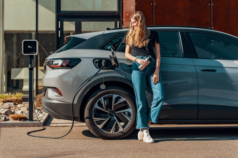 Imagem de uma moça recostada em um carro cinza que está sendo alimentado por uma tomada de eletricidade ilustra o artigo cujo título diz que uma abordagem inovadora na recarga de EVs pode acelerar sua adoção.