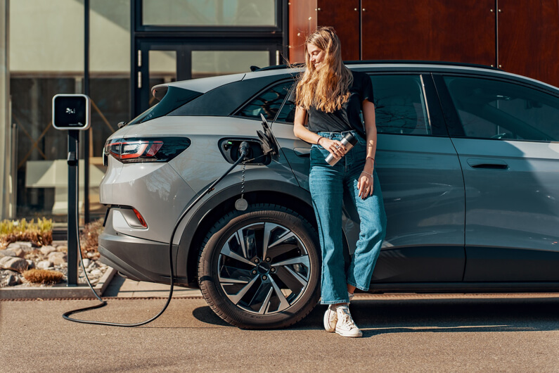 Imagem de uma moça recostada em um carro cinza que está sendo alimentado por uma tomada de eletricidade ilustra o artigo cujo título diz que uma abordagem inovadora na recarga de EVs pode acelerar sua adoção.